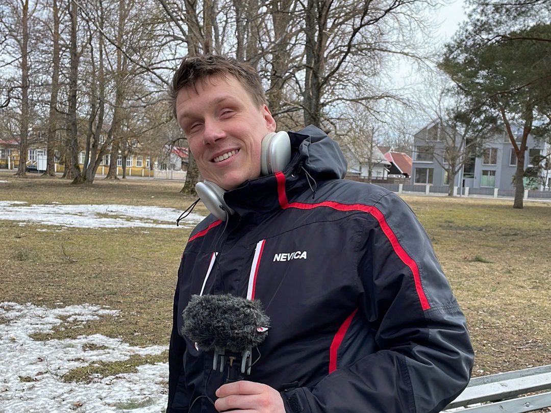 Jakob rosin standing in a park and holding a sound recorder, with a fluffy windscreen on top of it. He is wearing headphones.