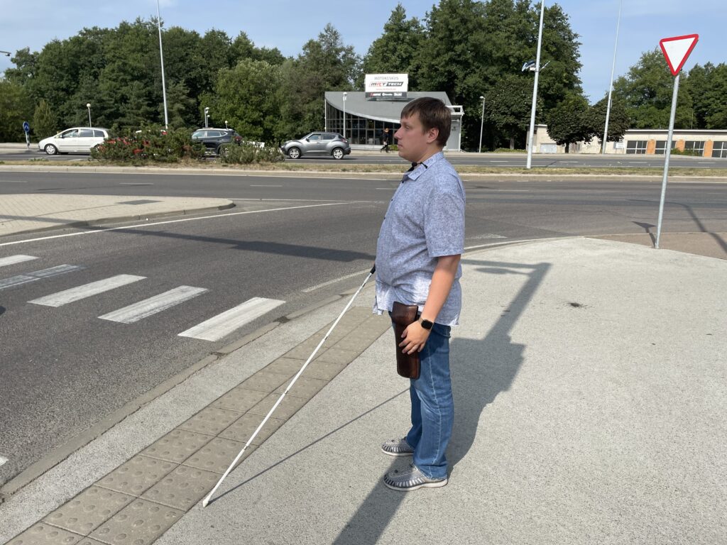 Jakob stands with a white cane in his hand at a city intersection, where tactile pavement can be seen on the ground in front of him.